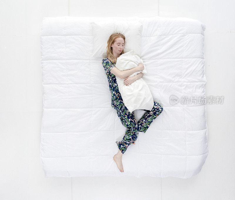 上图/全身/一个20-29岁的成年美丽的白人女性/年轻女性躺下/打盹/睡觉/放松/休息在卧室的白色背景下穿着睡衣拥抱
